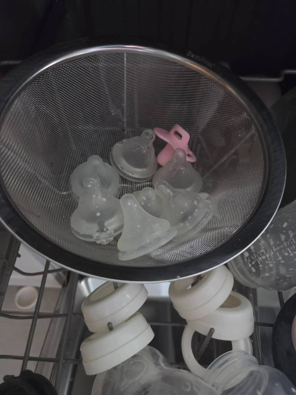 An open metal mesh colander holding bottle nipples and a pink pacifier on our dishwasher rack, with bottle parts below