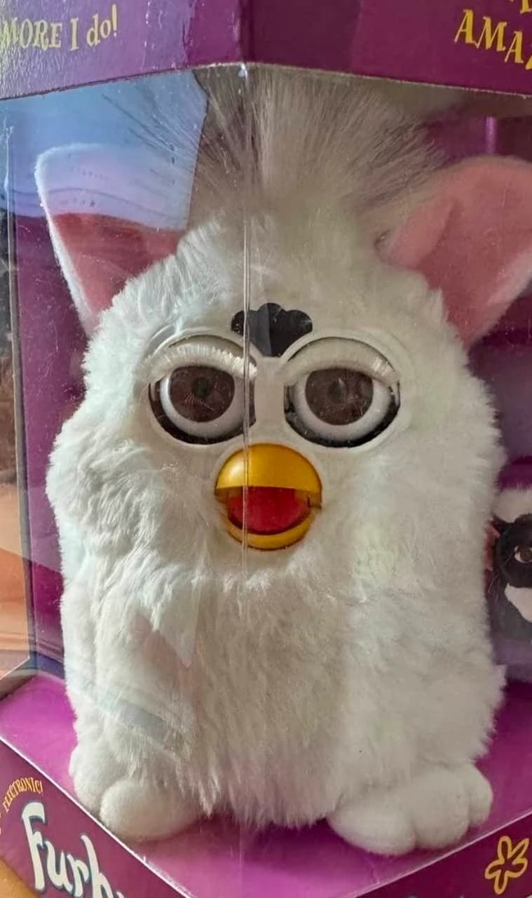 White Furby with pink ears and a yellow beak inside purple packaging