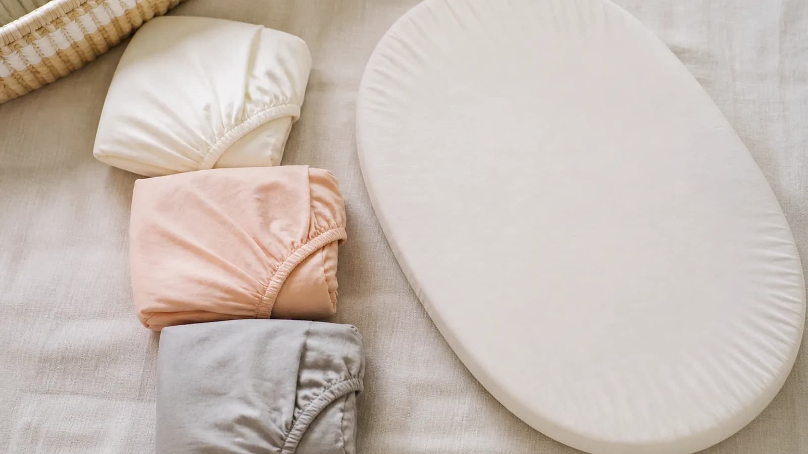 Ivory, peach, and gray fitted bassinet sheets arranged beside an oval mattress pad