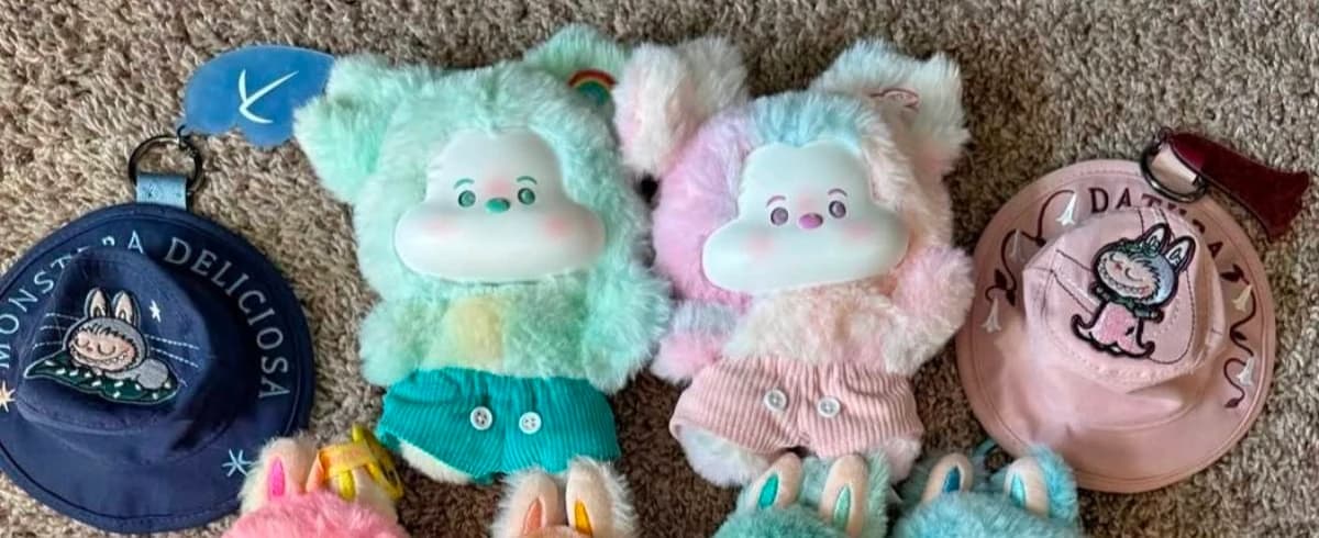 Two dressed plush dolls with teal and pink shorts beside miniature hats