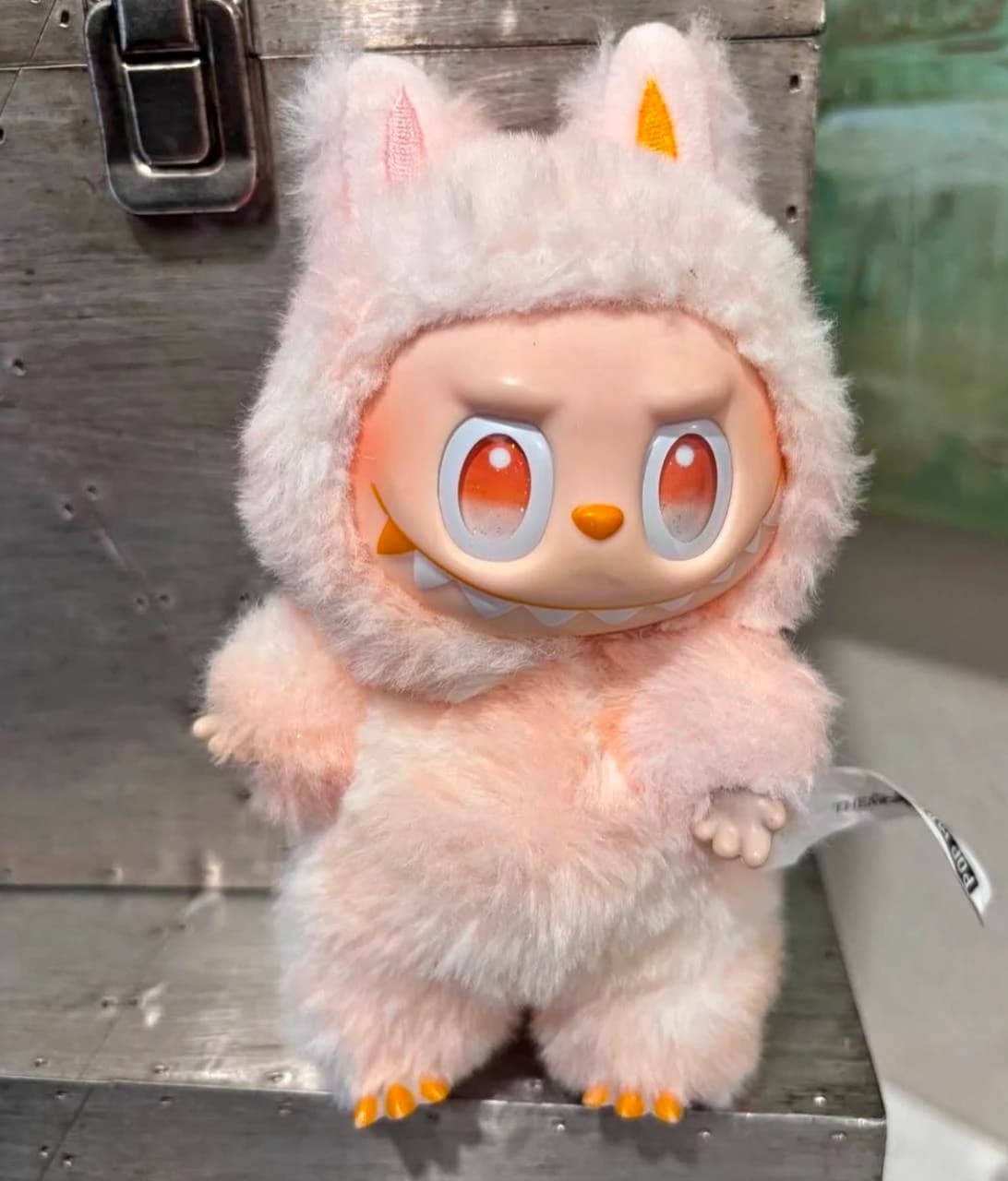 Pink-and-cream Labubu plush with orange eyes and an orange nose against wood