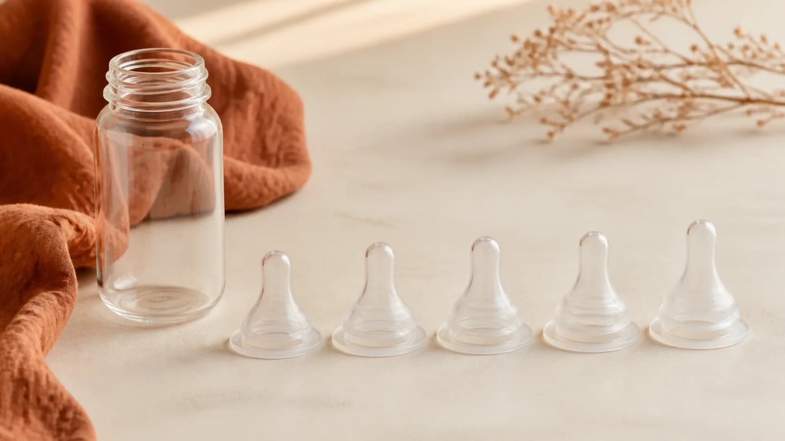 Five unbranded symmetrical silicone bottle nipples beside a clear bottle on a cream and rust surface
